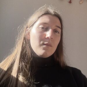 In a webcam headshot, central in the image is Alexandra with sunlight pouring in from the left side of the image frame, illuminating half of her
face. Alexandra is white, with light-brown hair and hazel eyes.